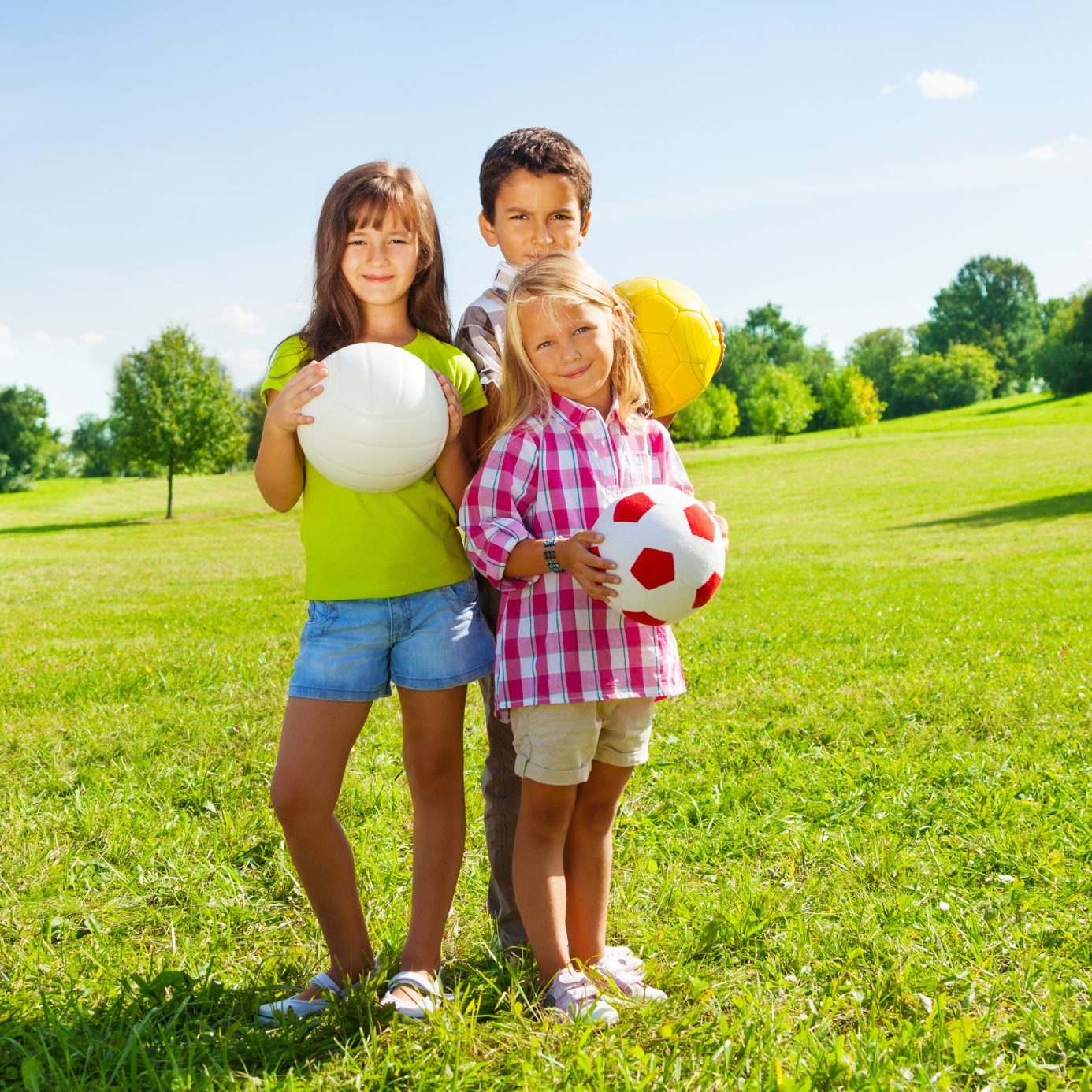 Three young kids holding sports balls in a grass field.