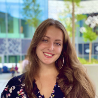 Young woman with long, wavy brown hair wearing a floral blouse and hoop earrings, smiling outdoors with modern buildings in the background.