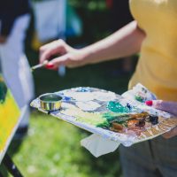Close-up of person painting outdoors, holding a palette with vivid paint colors and a brush, applying paint to a canvas on an easel.