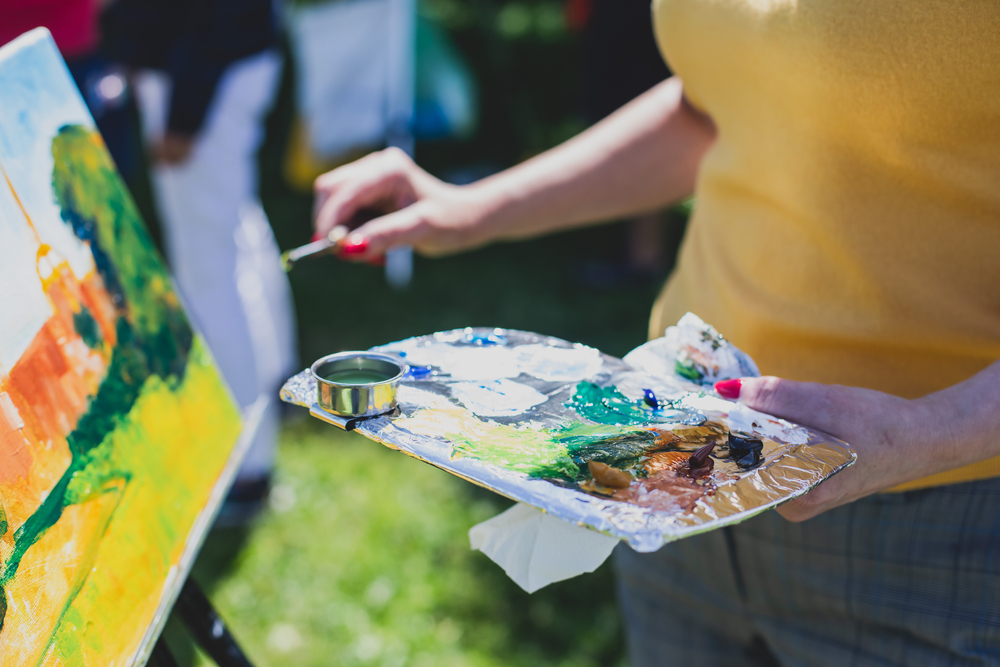 Close-up of person painting outdoors, holding a palette with vivid paint colors and a brush, applying paint to a canvas on an easel.