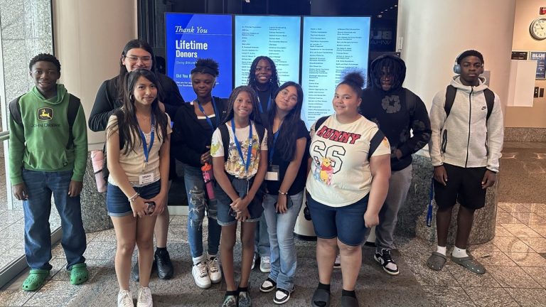 A group of nine young people stands in front of a display board titled "Thank You Lifetime Donors."