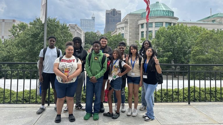 A group of ten people stands in front of a railing, with a city skyline and a domed building in the background.