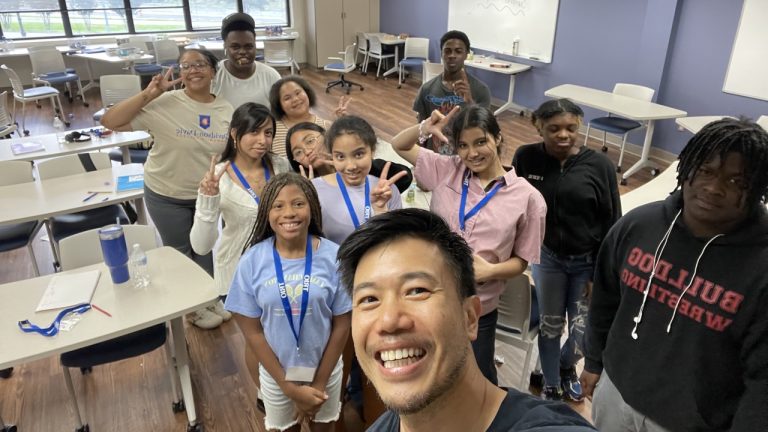 A group of people smiling and posing in a classroom with desks and large windows.