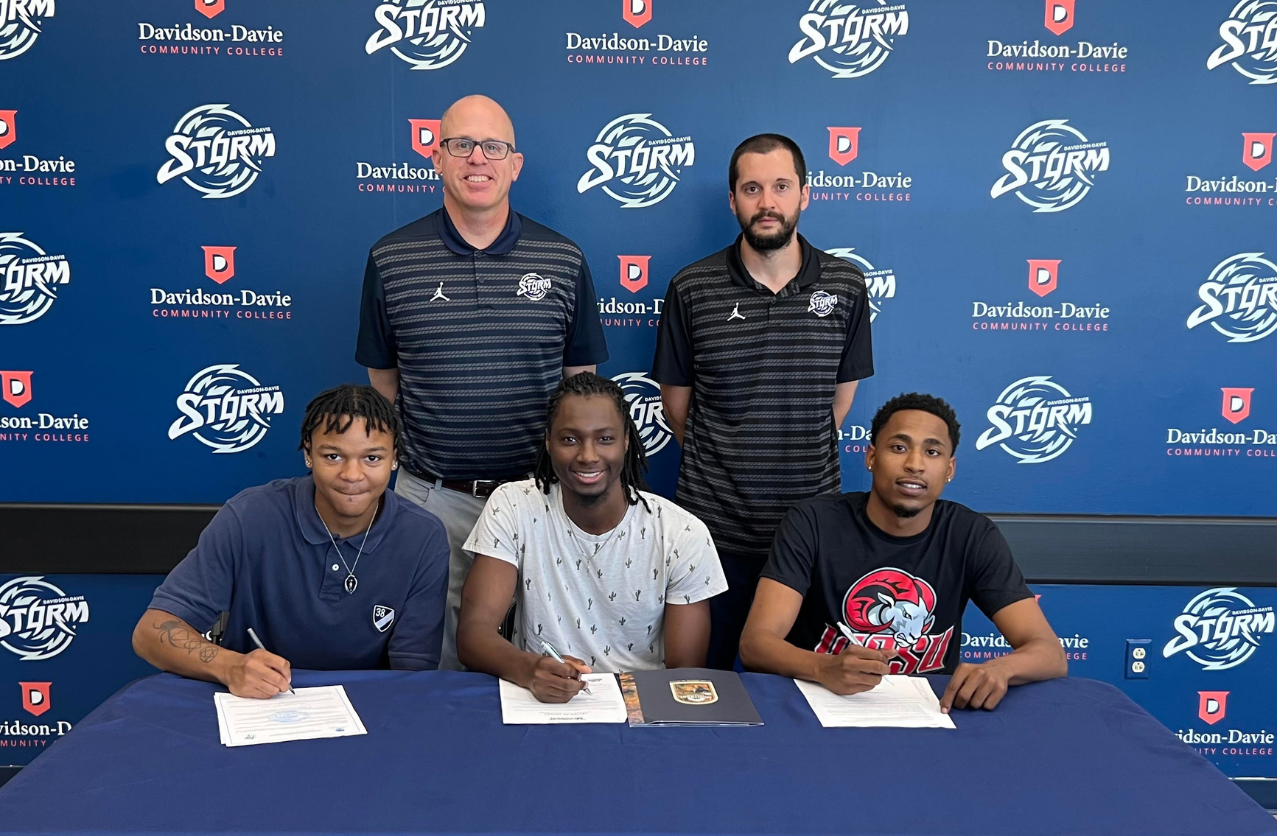 Three people signing documents at a table with two standing behind, all in front of a branded backdrop.