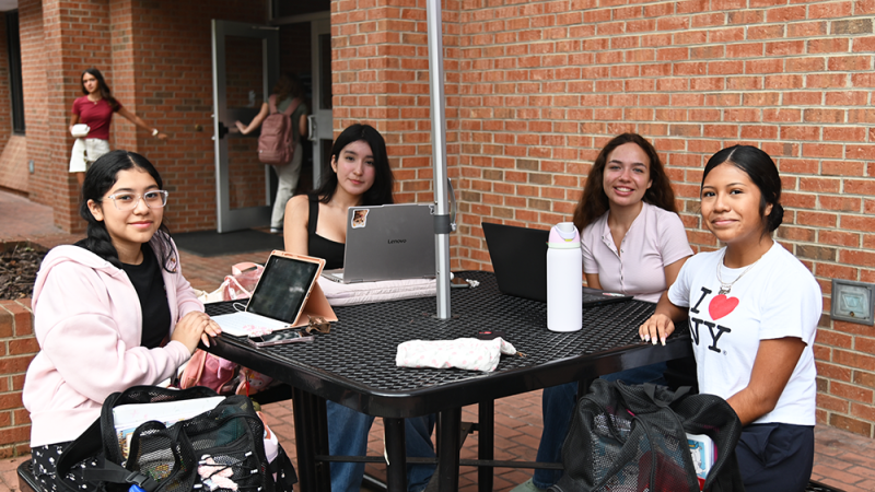 Four people sitting at a table with laptops and a tablet outside a brick building.
