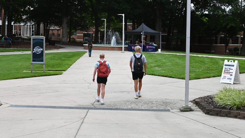 Two people walking on a campus path towards a fountain, with signs around them.