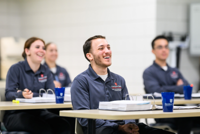 People in a classroom, wearing matching shirts, smiling and seated at tables with notebooks and binders.