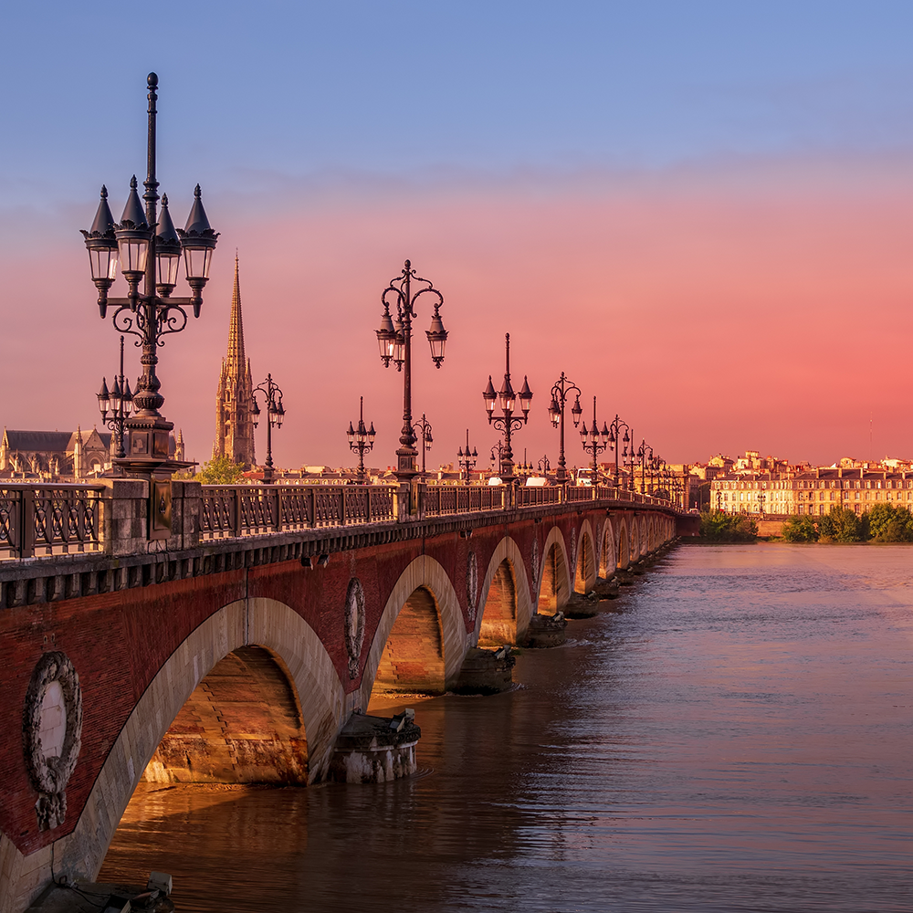 Stone bridge with arches and ornate lamps over a river at sunset, with a cityscape and church spire in the background.