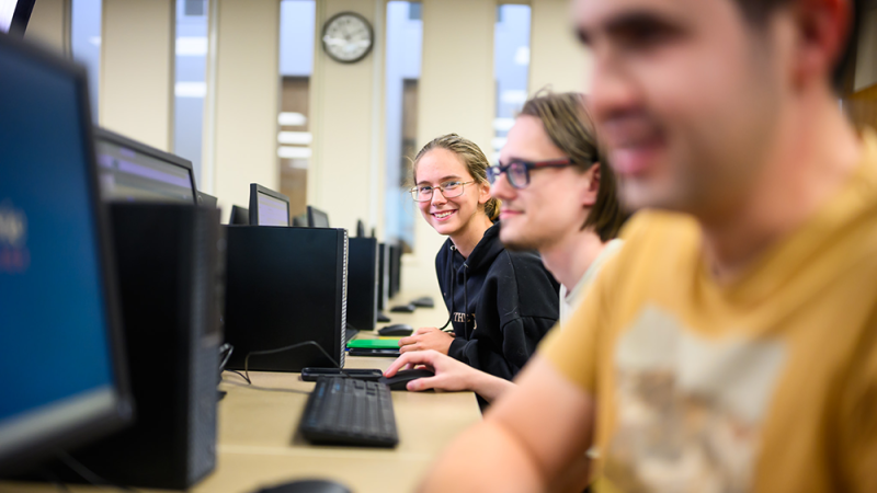 People working on computers in a lab, focusing on a smiling individual in a black hoodie.