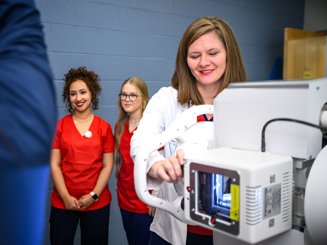 A woman in a white coat operates medical imaging equipment while two women in red scrubs observe.