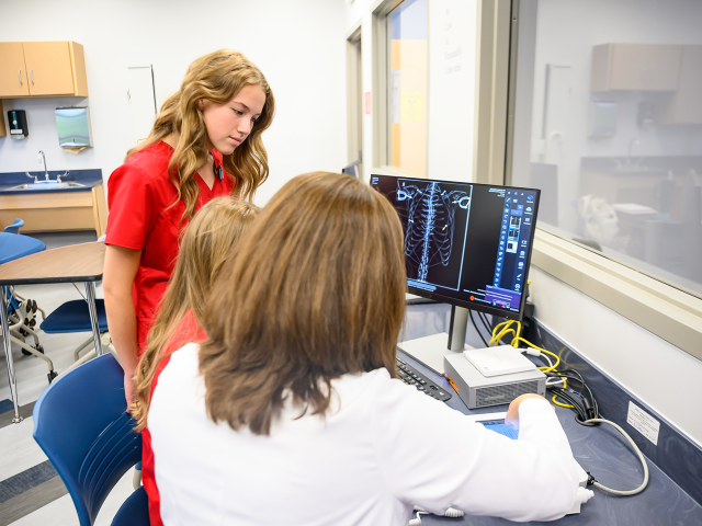 Three people examine an X-ray image of a rib cage on a computer monitor in a medical setting.