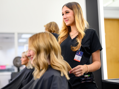 A hair stylist in a black uniform smiles while attending to a client with wavy blonde hair in a salon.