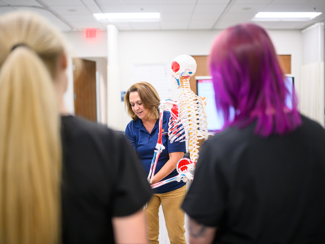 A person in a blue shirt stands beside a detailed skeleton model in a classroom, with two students listening.