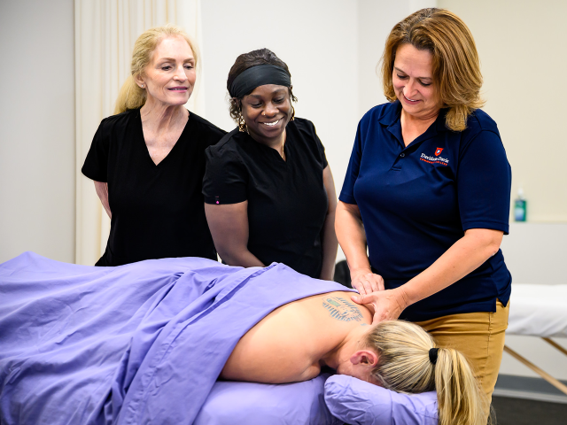A massage therapist gives a shoulder massage to a person lying on a table, observed by two others.